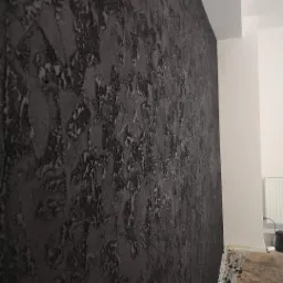 Dekoracyjna ściana w odcieniach szarości z wyraźną, nieregularną fakturą, widoczna pod kątem, z fragmentem białej ściany i grzejnika w tle.