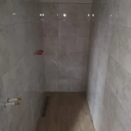 Wnętrze kabiny prysznicowej w trakcie wykańczania: jasne, marmurkowe płytki ścienne, drewnopodobne płytki podłogowe, widoczne rury instalacji wodnej i poziomica.