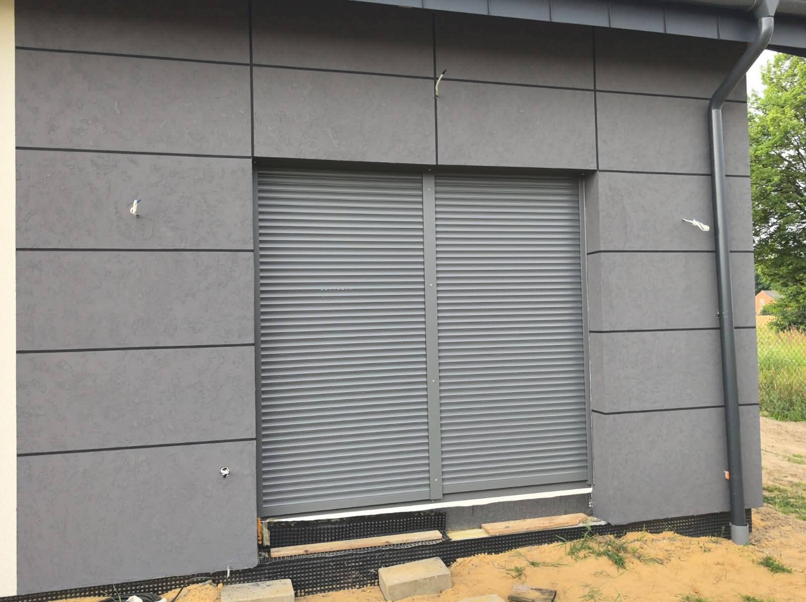 Szara elewacja budynku z panelami i roletami zewnętrznymi. Widoczne detale wykończenia, elementy montażowe i fragment krajobrazu. Ujęcie z bliska.
