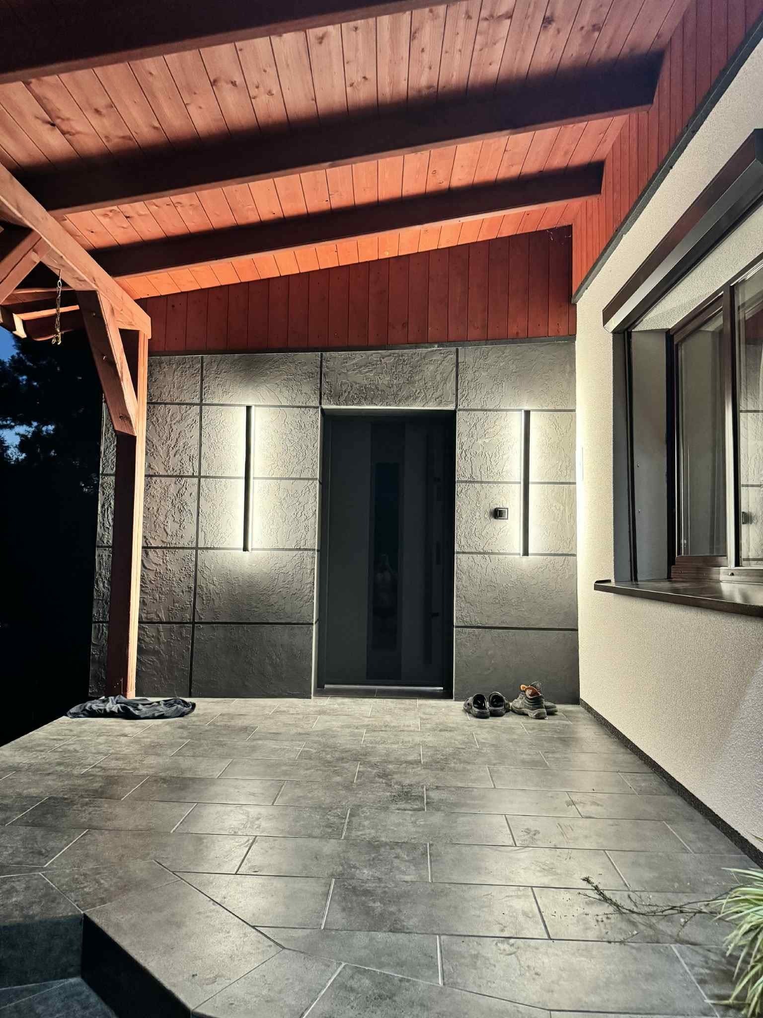 Nowoczesna elewacja domu z szarymi płytami i oświetleniem LED. Ciemna podłoga z płytek, drewniany podbitka dachu. Widoczne buty przed wejściem.