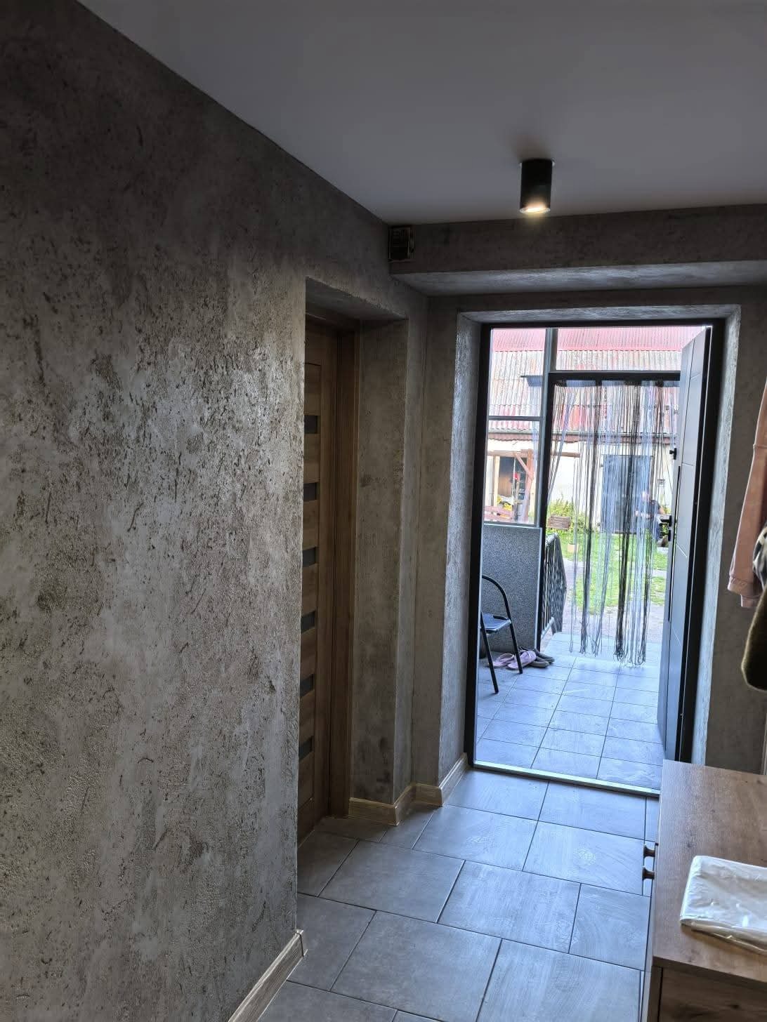 Szara ściana z efektem dekoracyjnym, płytki podłogowe w odcieniach szarości i widok na zewnątrz przez otwarte drzwi wejściowe. Nowoczesne wykończenie wnętrza.