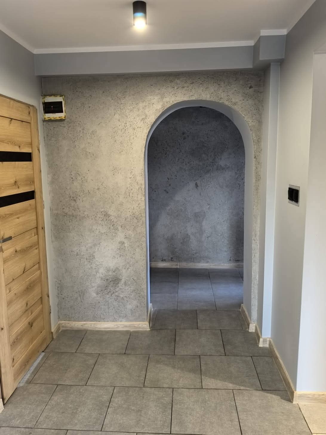 Wykończony korytarz z łukowatym przejściem i szarą, teksturowaną ścianą. Na podłodze szare płytki. Drewniane drzwi z czarnymi akcentami po lewej stronie.