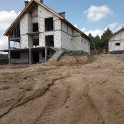 Budowa domu murowanego z betonu komórkowego w stanie surowym otwartym, widoczne ściany, schody zewnętrzne i częściowo wykonany dach, na drugim planie mniejszy budynek w podobnym stanie.