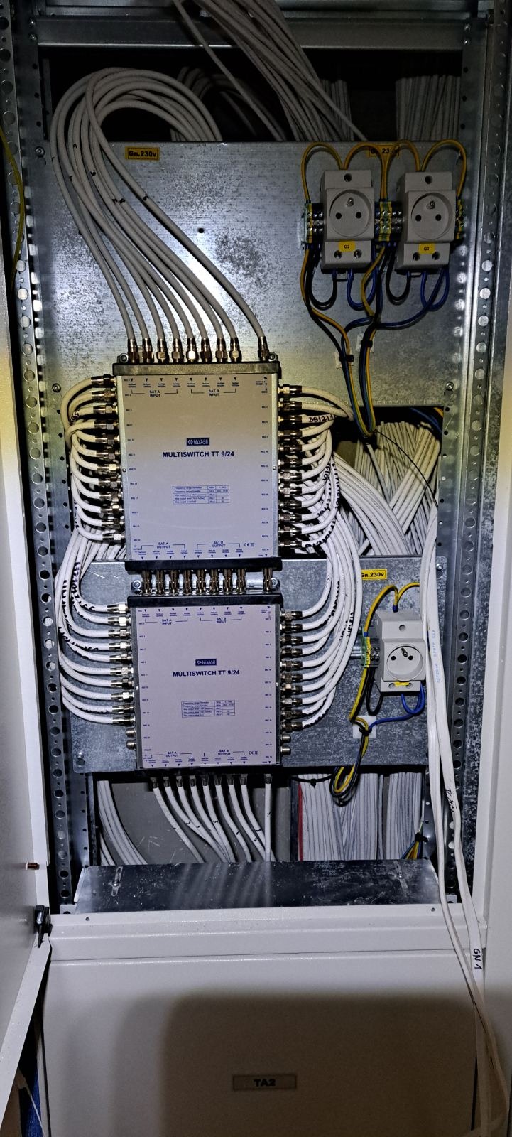 Rozdzielnia elektryczna z dwoma multiswitchami TT 9/24, licznymi kablami koncentrycznymi i gniazdkami 230V w metalowej szafie rack.