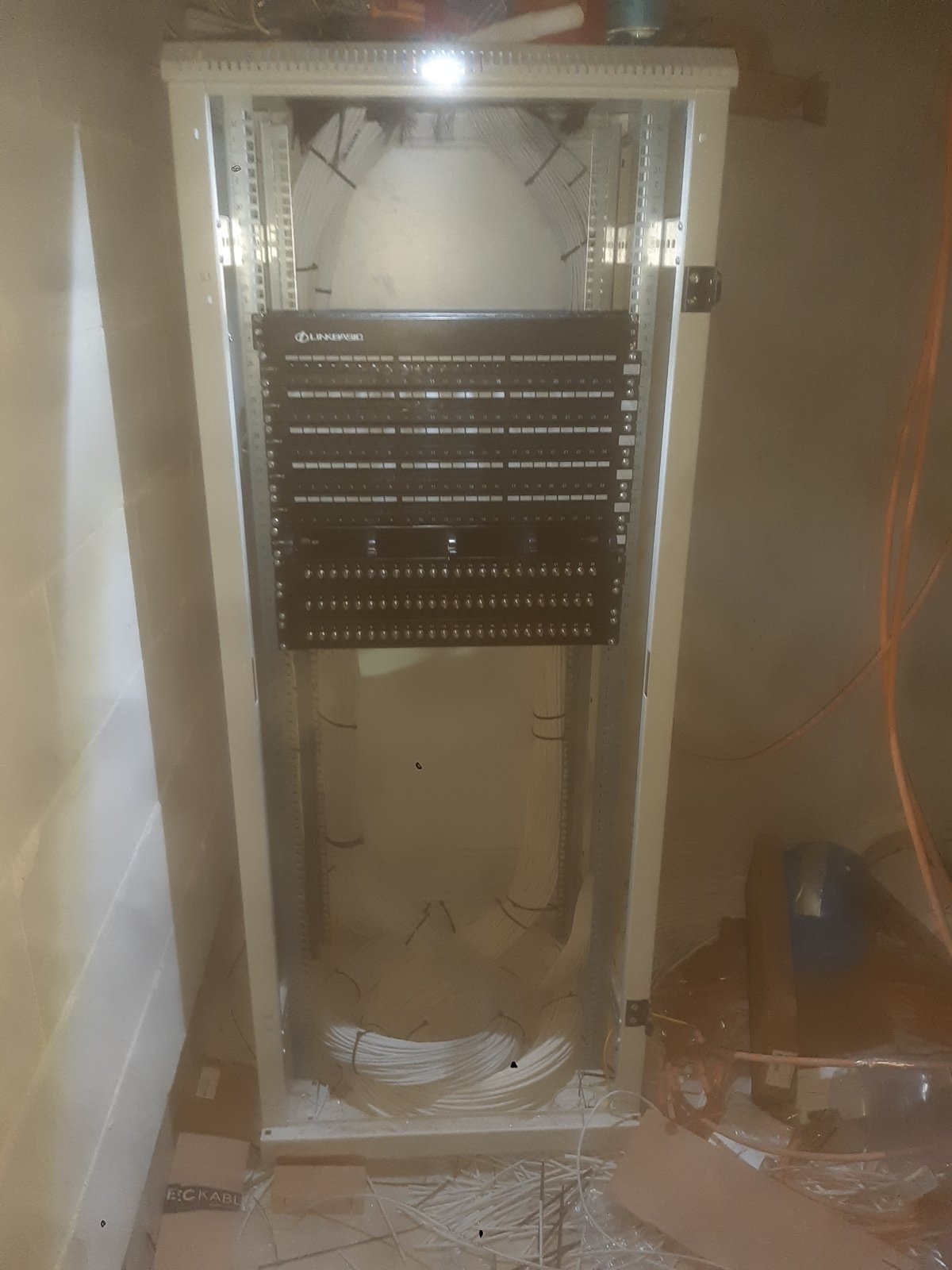 Panel krosowniczy Linkbasic zamontowany w szafie rack z uporządkowanymi kablami, widoczne okablowanie strukturalne, ściana z bloków betonowych w tle.