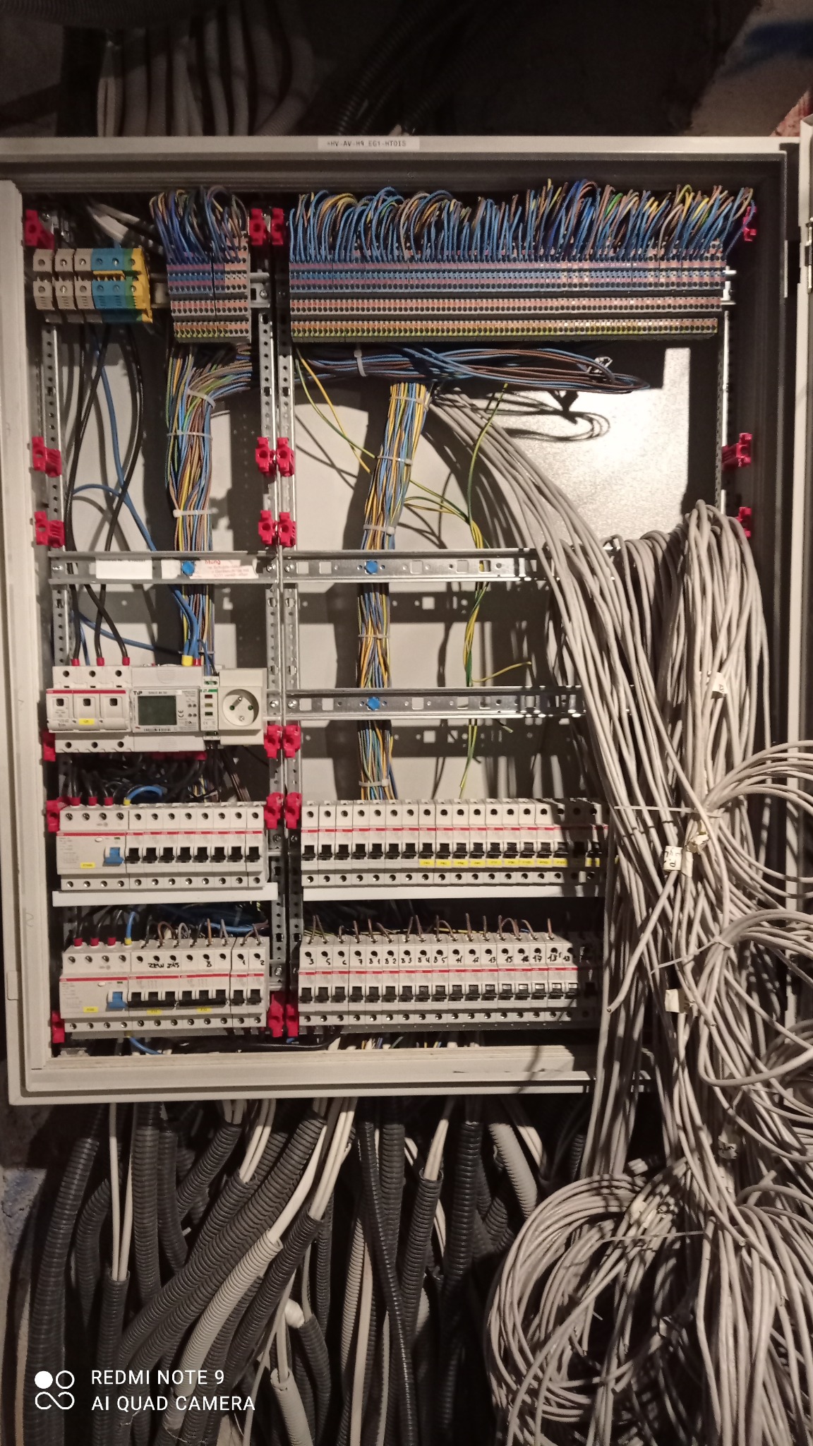 Otwarta szafa elektryczna z zamontowanymi bezpiecznikami, przewodami w różnych kolorach i grubościach, częściowo ułożonymi w peszlach, widoczne elementy montażowe na szynach DIN.