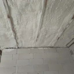 Poddasze ocieplone pianką PUR, widoczne krokwie i ściana z bloczków, pianka o nieregularnej strukturze pokrywa całą powierzchnię.