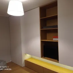 Biała zabudowa meblowa z drewnianymi akcentami, obejmująca szafę, otwarte półki i niską komodę z podświetleniem LED, w pokoju z drewnianą podłogą i widocznym fragmentem dziecięcego łóżka.