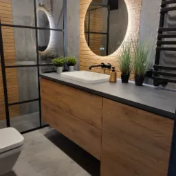 Nowoczesna łazienka z drewnianymi akcentami: szafka pod umywalkę z blatem w kolorze grafitowym, okrągłe lustro z podświetleniem LED, kabina prysznicowa z czarnymi profilami i grzejnik drabinkowy...