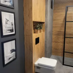 Nowoczesna toaleta podwieszana z zabudową z drewna w odcieniach brązu, z półką na rośliny w plecionym koszyku, na ścianie czarno-białe zdjęcia w czarnych ramkach, szara podłoga i ściany, drewniana...