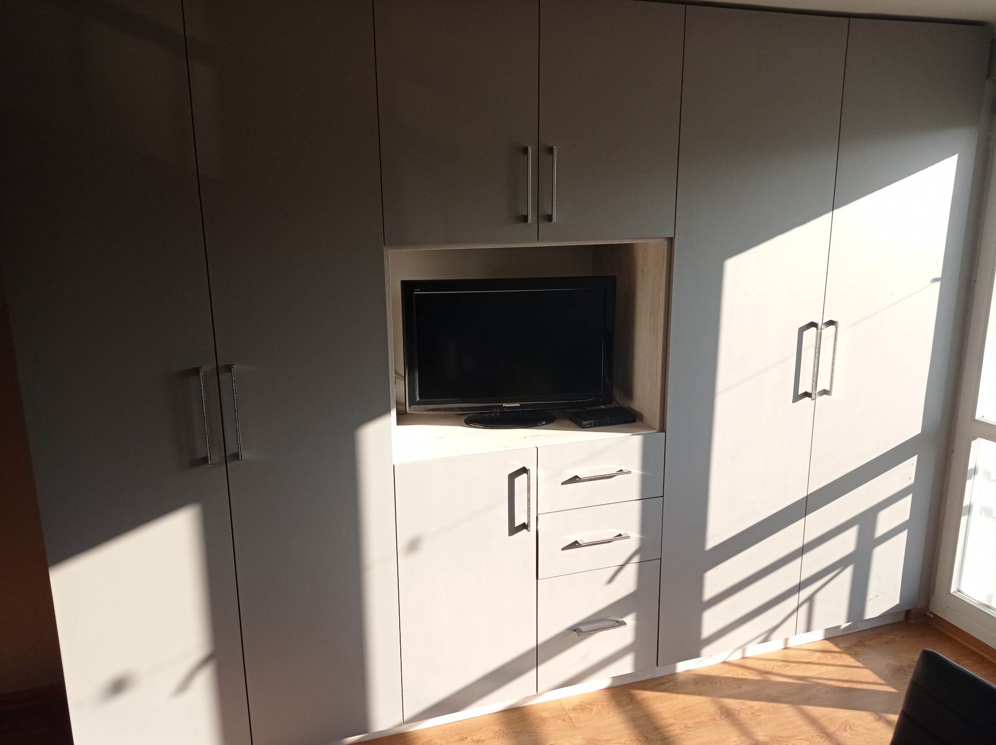 Jasnoszara szafa wnękowa z wbudowanym miejscem na telewizor, oświetlona promieniami słonecznymi padającymi przez okno.