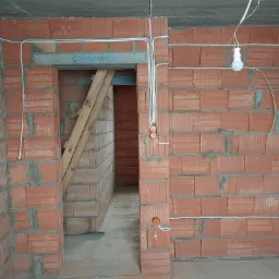 Surowy stan budynku w trakcie prac: widoczne ceglane ściany z poprowadzonymi przewodami elektrycznymi, tymczasowe oświetlenie żarówką oraz drewniane stemple podtrzymujące nadproże.