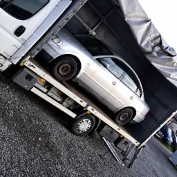 Srebrny samochód osobowy zabezpieczony w przestrzeni ładunkowej samochodu dostawczego z plandeką, widoczne elementy mocowania ładunku.