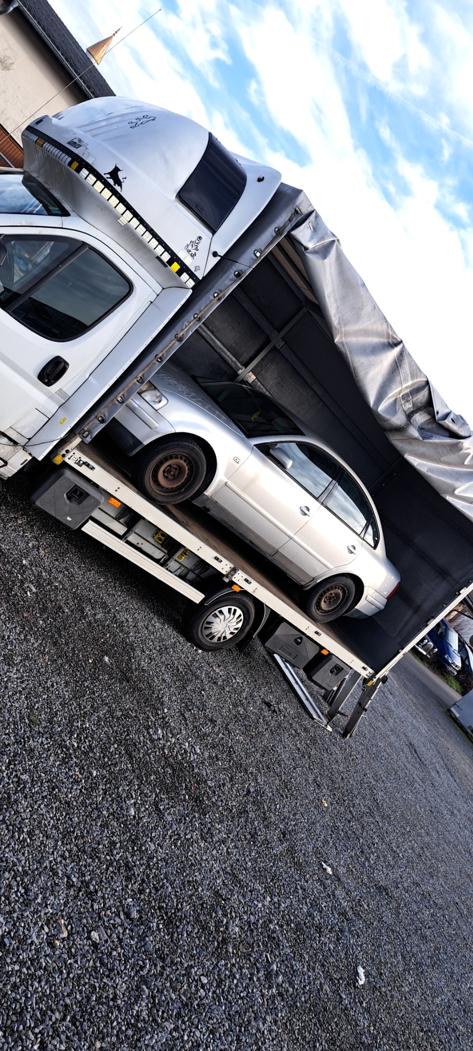 Srebrny samochód osobowy zabezpieczony w przestrzeni ładunkowej samochodu dostawczego z plandeką, widoczne elementy mocowania ładunku.