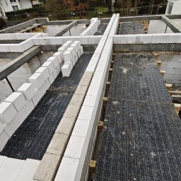 BOEK usługi budowlane Eduard Krynitski - Wysoki kąt na fundamenty domu z białych bloczków, częściowo ułożonych, z czarną siatką zbrojeniową wewnątrz. Widoczne drewniane podpory i fragment budynku w tle.