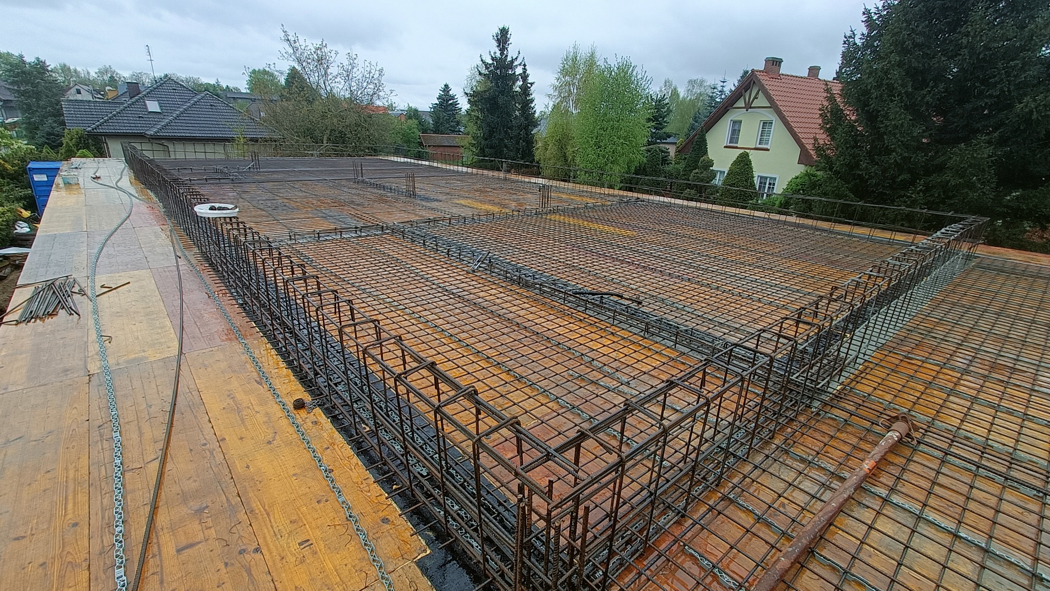 Siatka zbrojeniowa na deskowaniu szalunkowym, przygotowana do zalania betonem, widok z góry na tle zabudowy jednorodzinnej w pochmurny dzień.