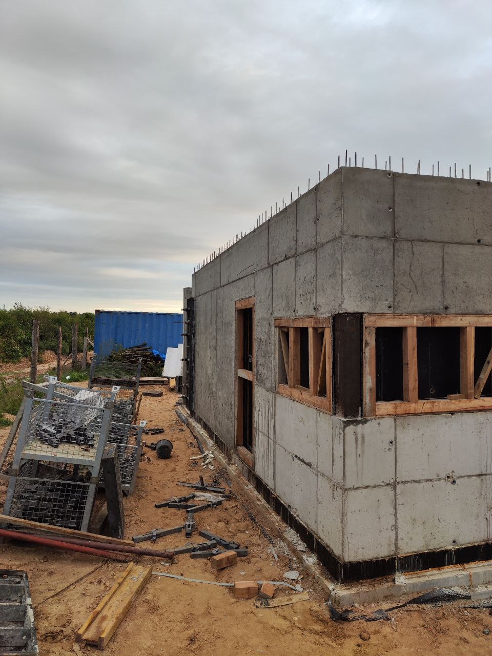 Surowy budynek żelbetowy w trakcie budowy. Widoczne szalunki okienne, wystające pręty zbrojeniowe i bałagan na placu budowy. W tle kontener.