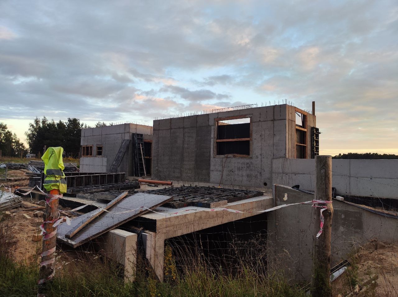 Budowa domu z betonu: surowe ściany zbrojone, widoczna konstrukcja, żółta kamizelka odblaskowa, szalunki okienne i zbrojenia na tle pochmurnego nieba.