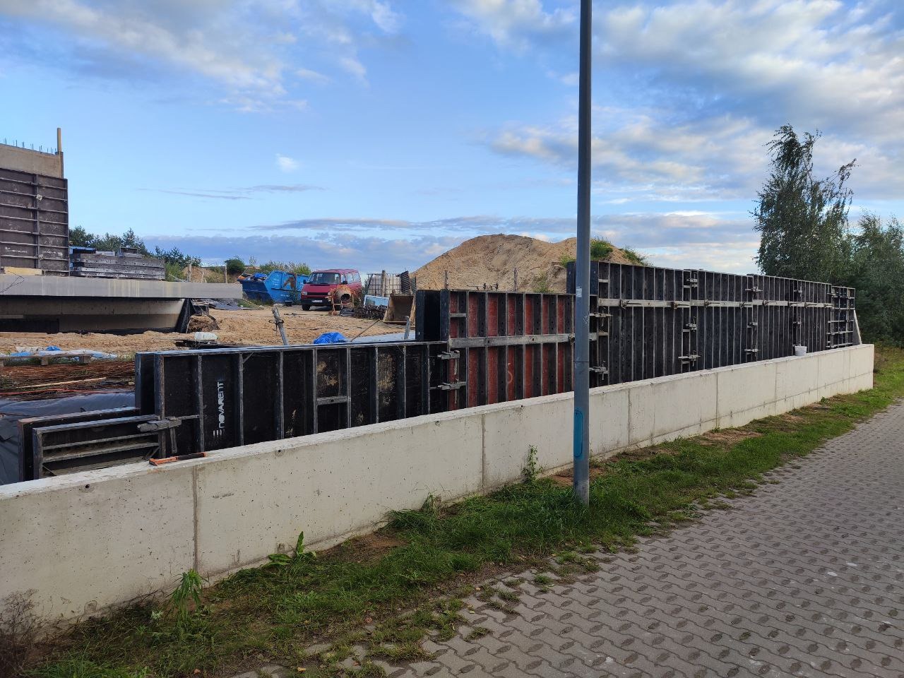 Szare betonowe fundamenty z czarnym szalunkiem, widok z boku na placu budowy z piaskiem i pojazdami w tle, w dziennym świetle.