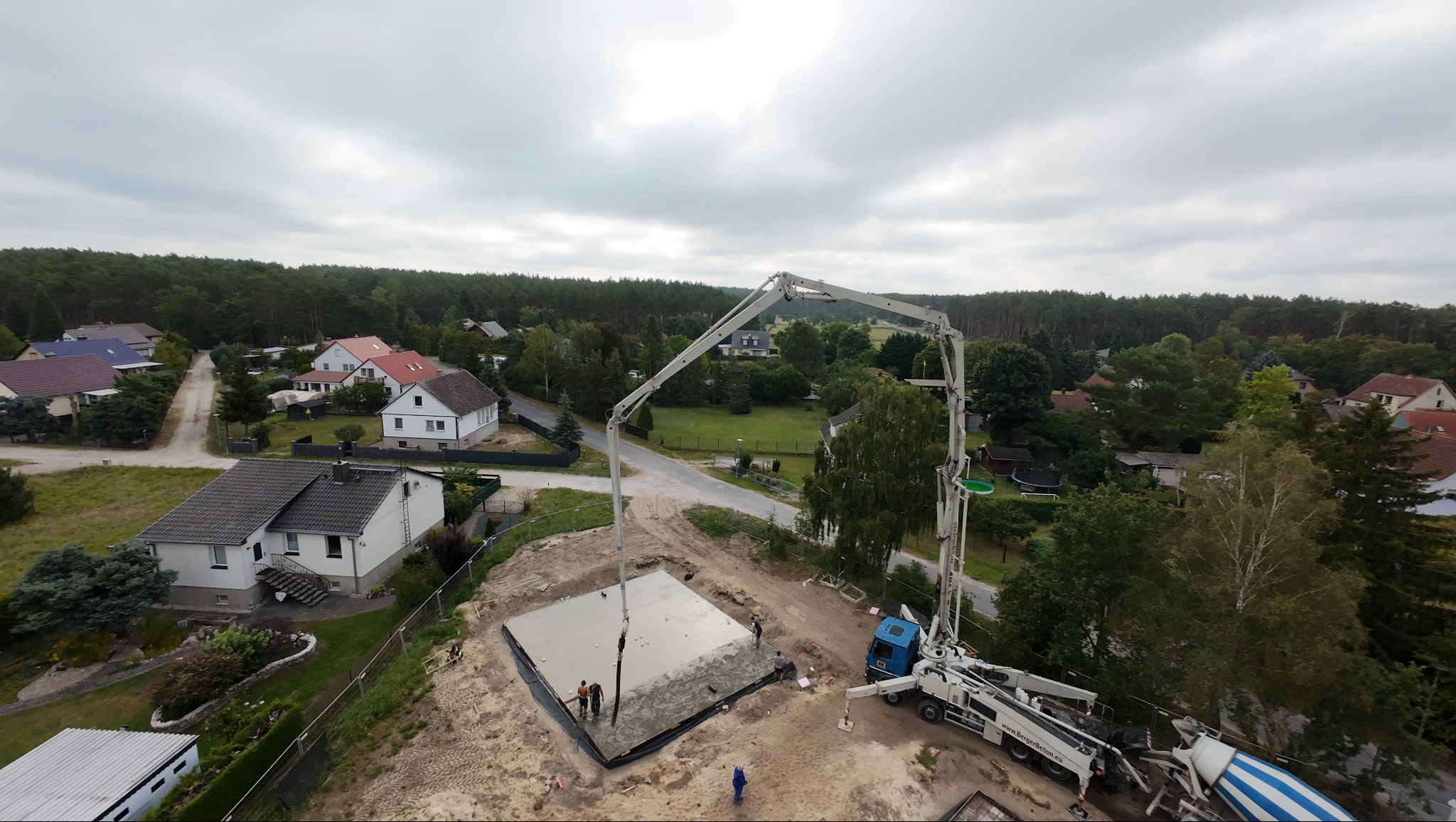 Betonowanie fundamentu domu jednorodzinnego z użyciem pompy do betonu. Widok z góry na plac budowy w otoczeniu zabudowy mieszkalnej i lasu, pochmurne niebo.
