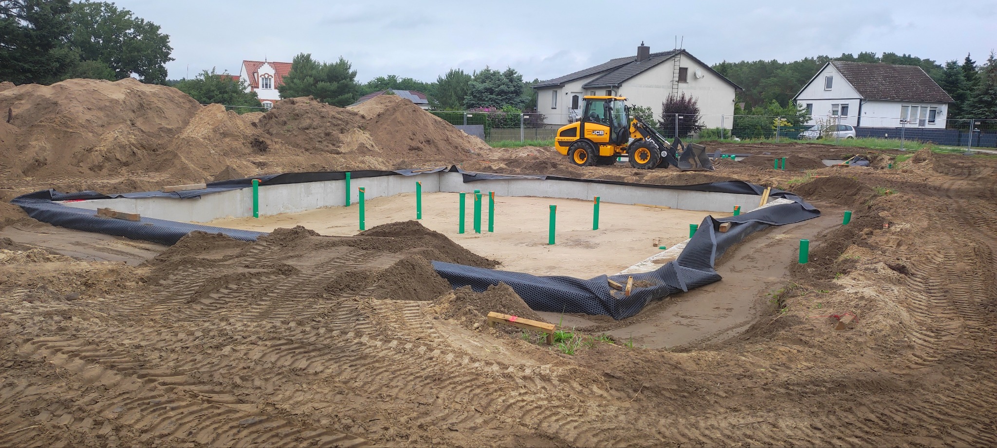 Fundamenty domu w trakcie budowy, widoczny wykop, izolacja, rury pionowe i koparka JCB w tle. Teren przygotowany pod wylewkę.