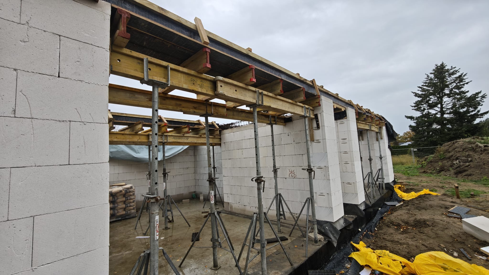 Wznoszenie ścian z białych bloczków, wspartych metalowymi stemplami. Widoczne drewniane szalunki stropowe i worki z zaprawą. Tło: pochmurne niebo i drzewo iglaste.