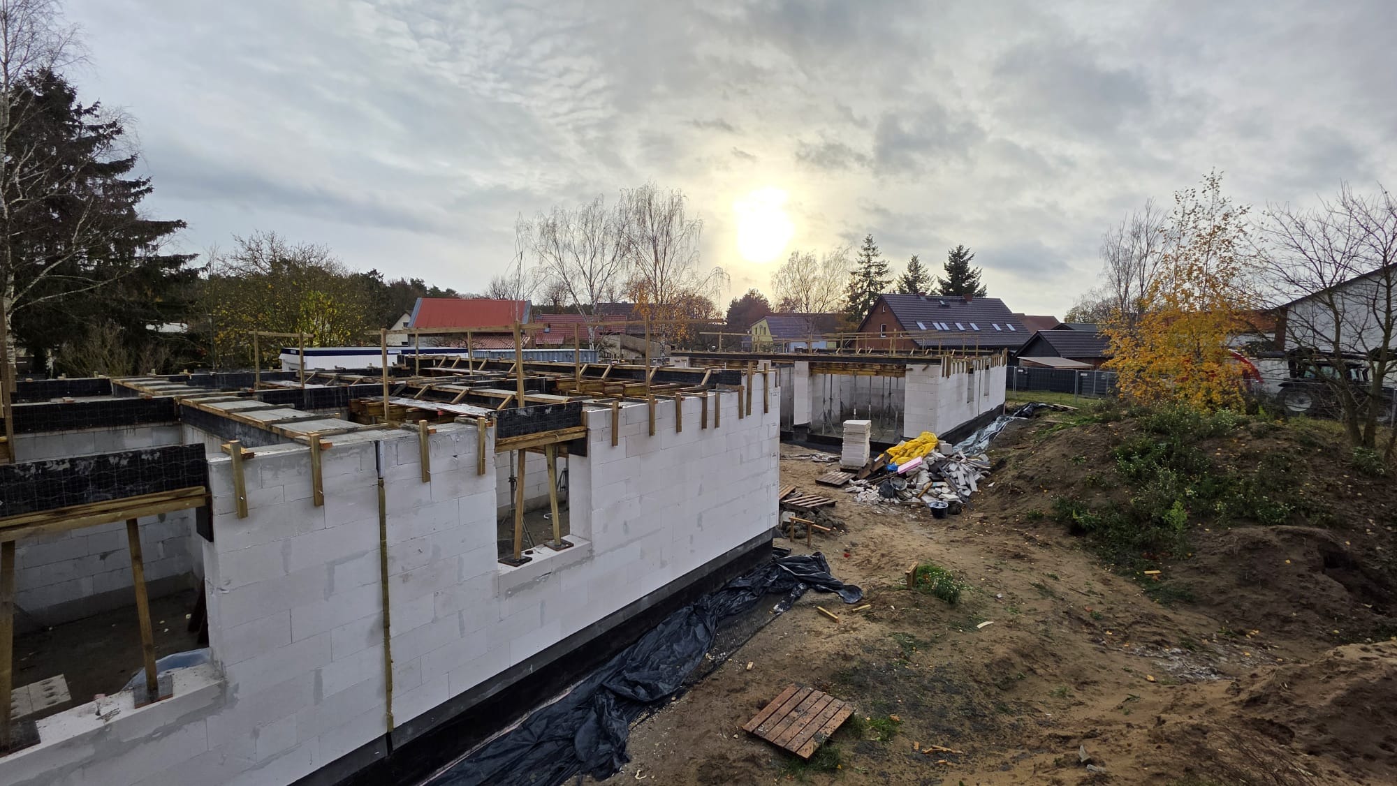 Wznoszenie ścian parteru domu z betonu komórkowego, widoczne szalunki i zbrojenie pod wieniec. W tle zabudowa jednorodzinna i pochmurne niebo.