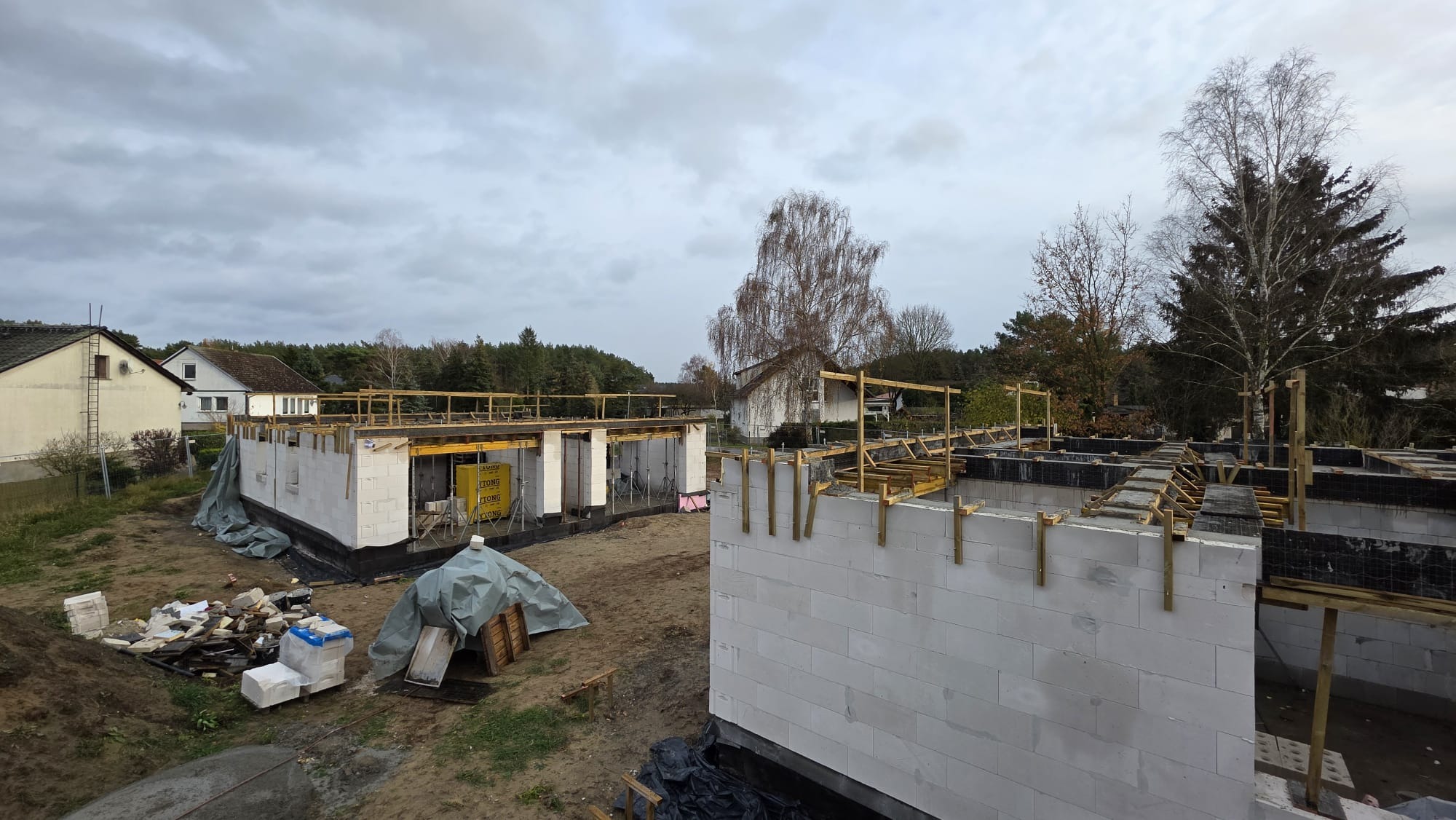 Budowa domu w toku: ściany z białych bloczków, drewniane szalunki, widok z góry na plac budowy w pochmurny dzień. Częściowo przykryte materiały budowlane.
