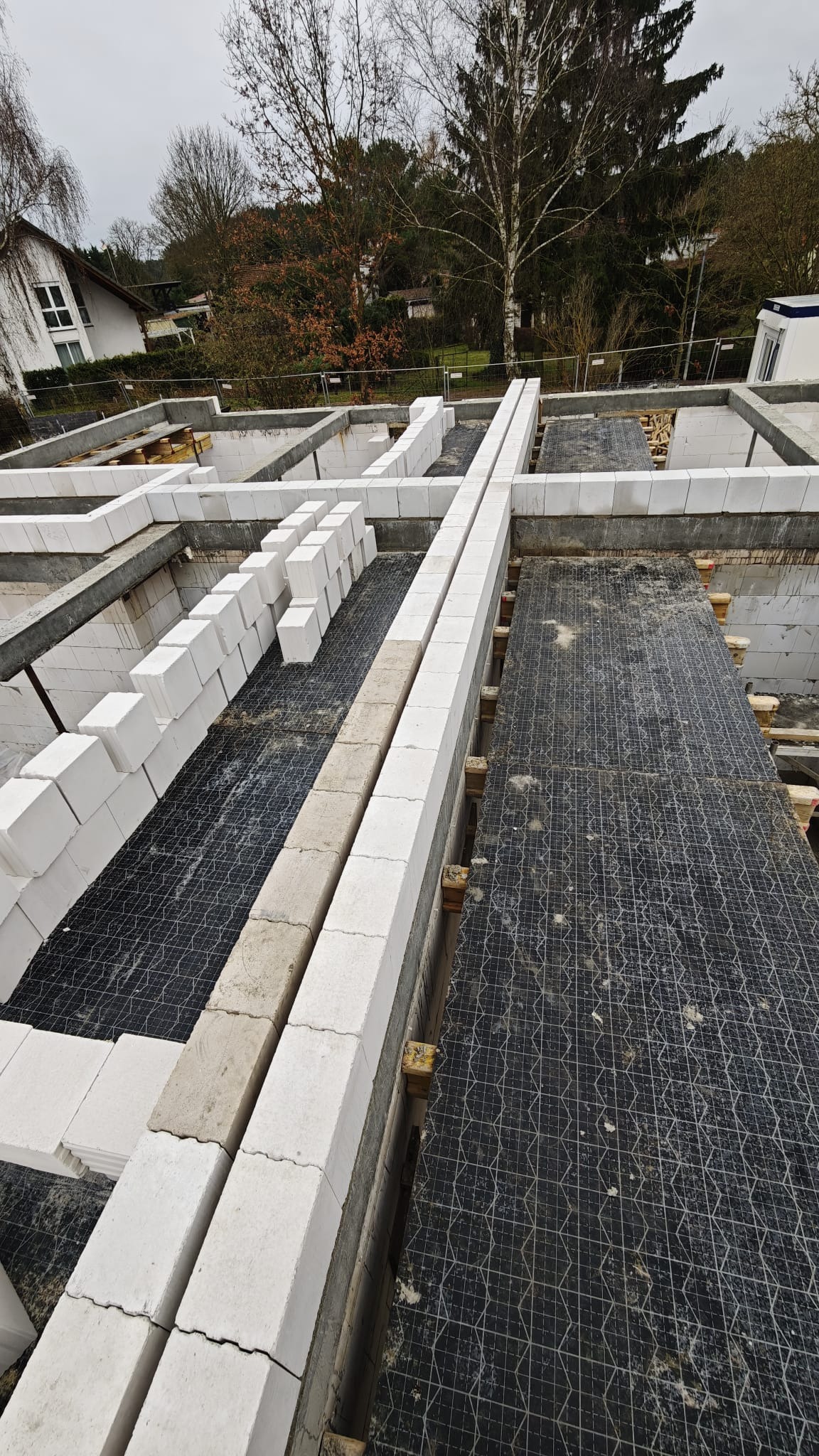 Wysoki kąt na fundamenty domu z białych bloczków, częściowo ułożonych, z czarną siatką zbrojeniową wewnątrz. Widoczne drewniane podpory i fragment budynku w tle.
