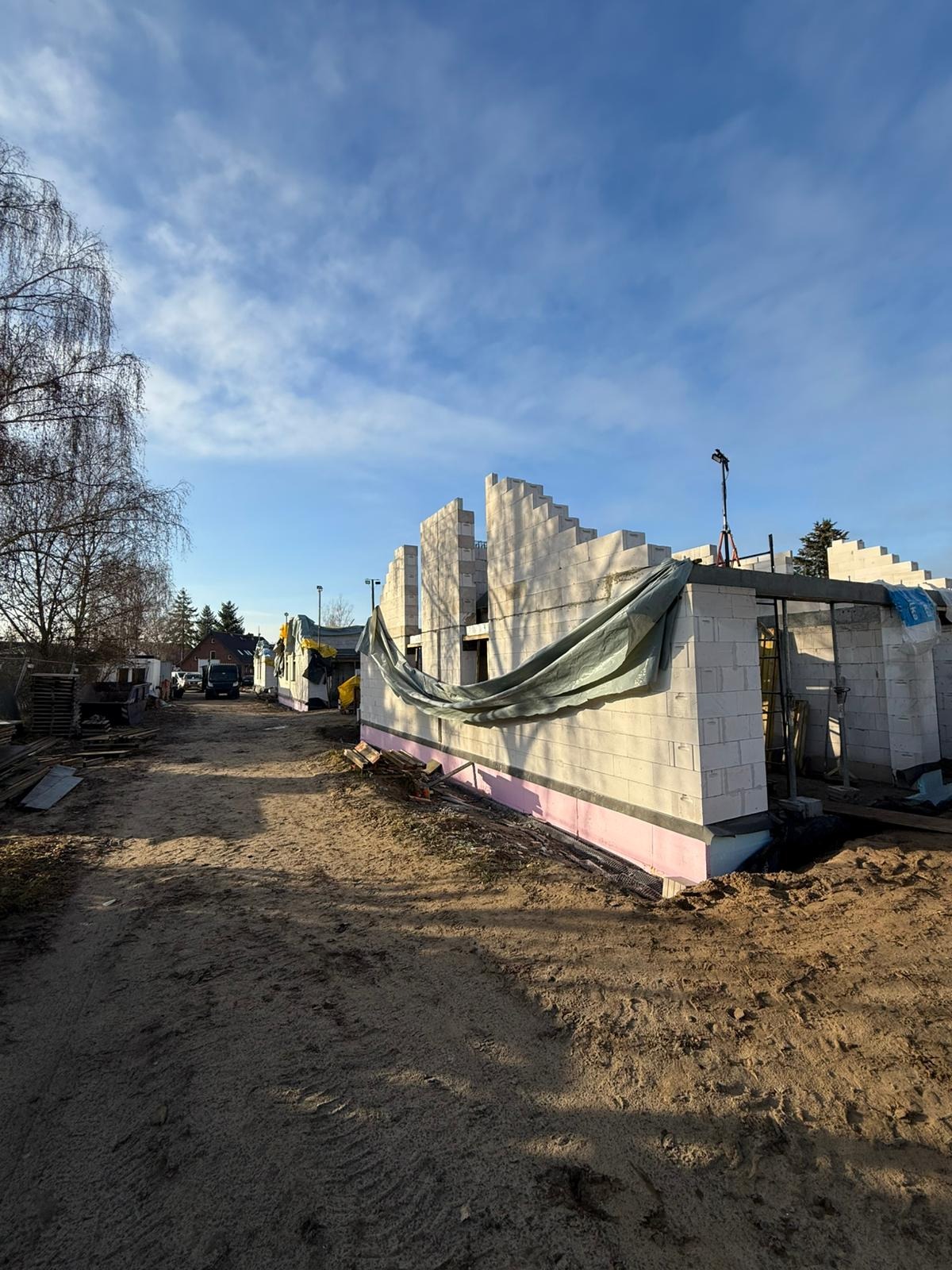 Wznoszenie ścian z bloczków betonu komórkowego na fundamencie z izolacją termiczną. Widoczne zbrojenia i częściowe przykrycie folią budowlaną. Budowa w toku, w słoneczny dzień.