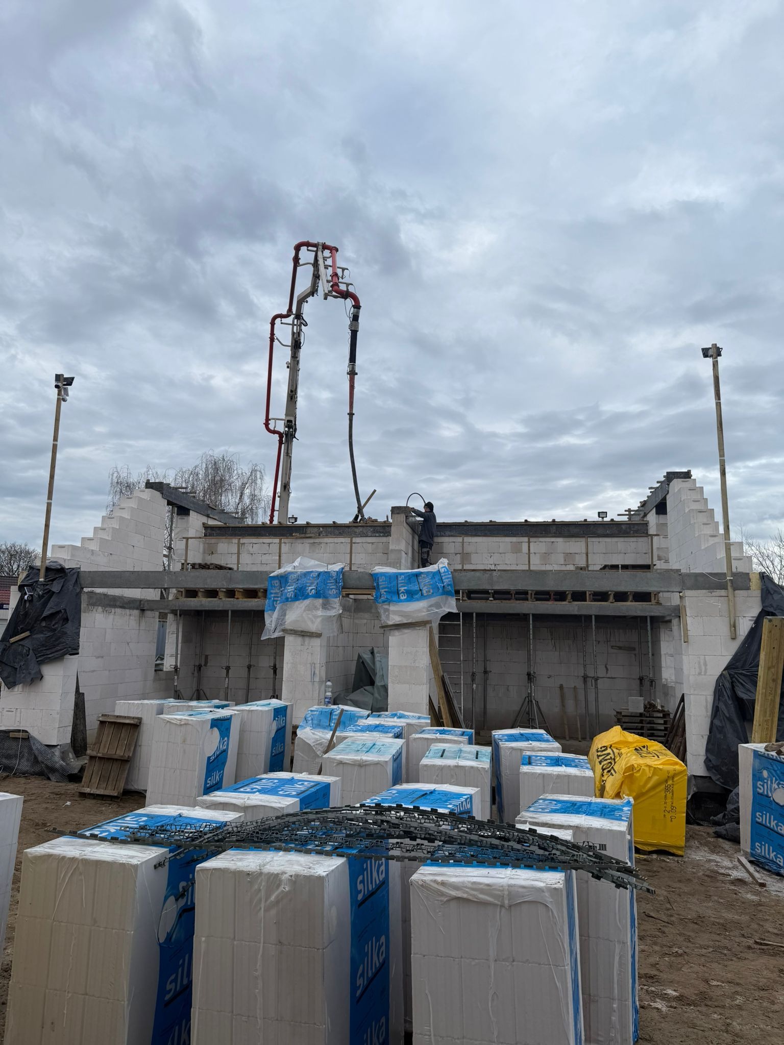 Budowa domu z betonu komórkowego Silka: ściany wzniesione, widoczna pompa do betonu, zbrojenie i materiały budowlane na placu budowy pod pochmurnym niebem.