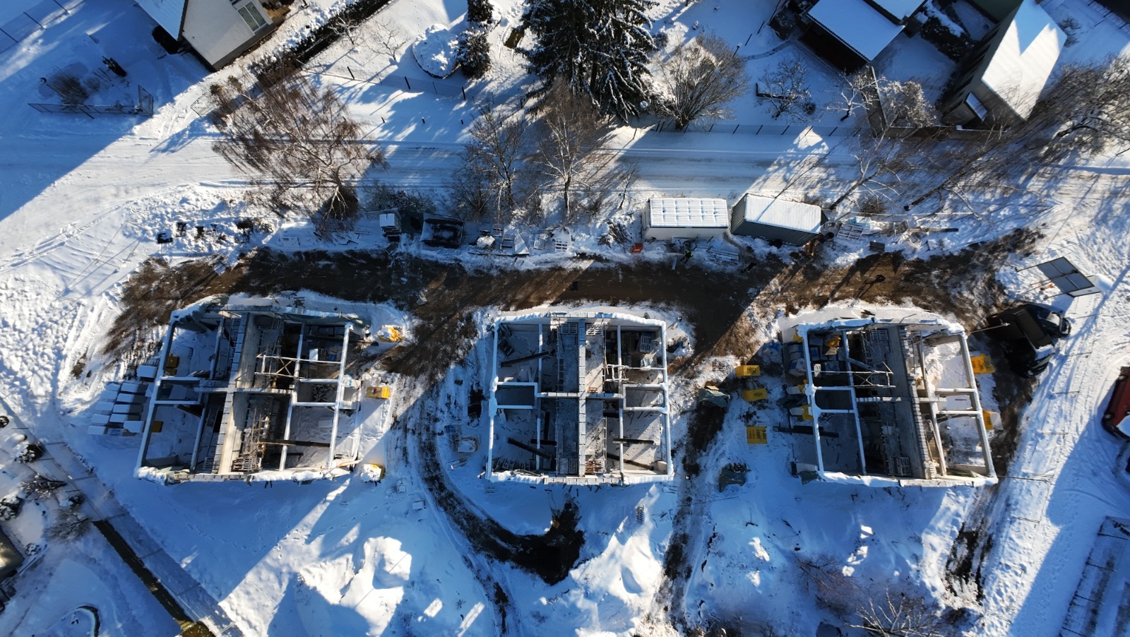 Zimowy widok z lotu ptaka na trzy budowy domów w stanie surowym otwartym, otoczone śniegiem i drzewami. Widoczne fundamenty i ściany nośne.