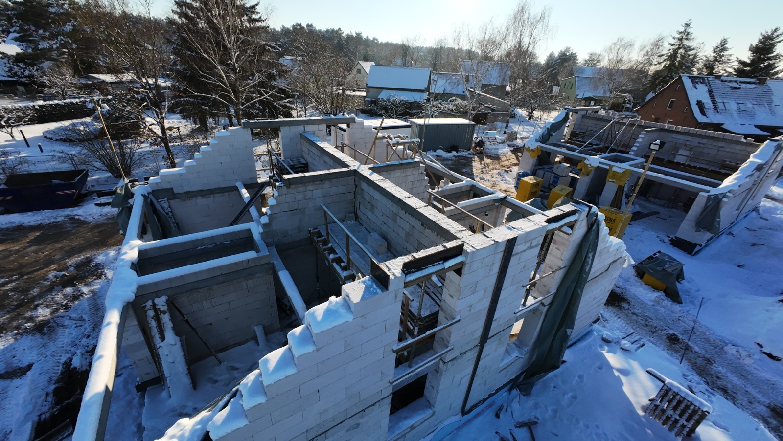 Wysoki kąt na budowę domu w zimie: białe bloczki ścian wznoszą się spod śniegu, drewniane belki stropowe, w tle zasypane śniegiem domy i drzewa.