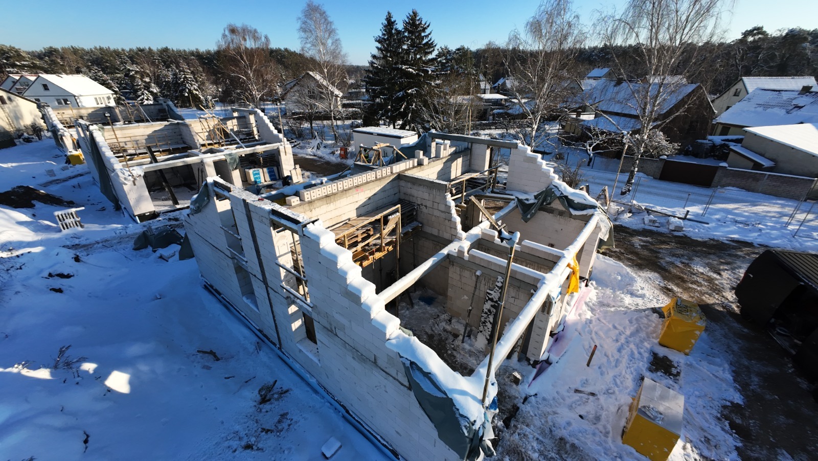 Stan surowy otwarty domu z białego bloczka w zimowej scenerii. Widok z góry ukazuje ściany bez dachu, drewniane szalunki i śnieżne otoczenie.