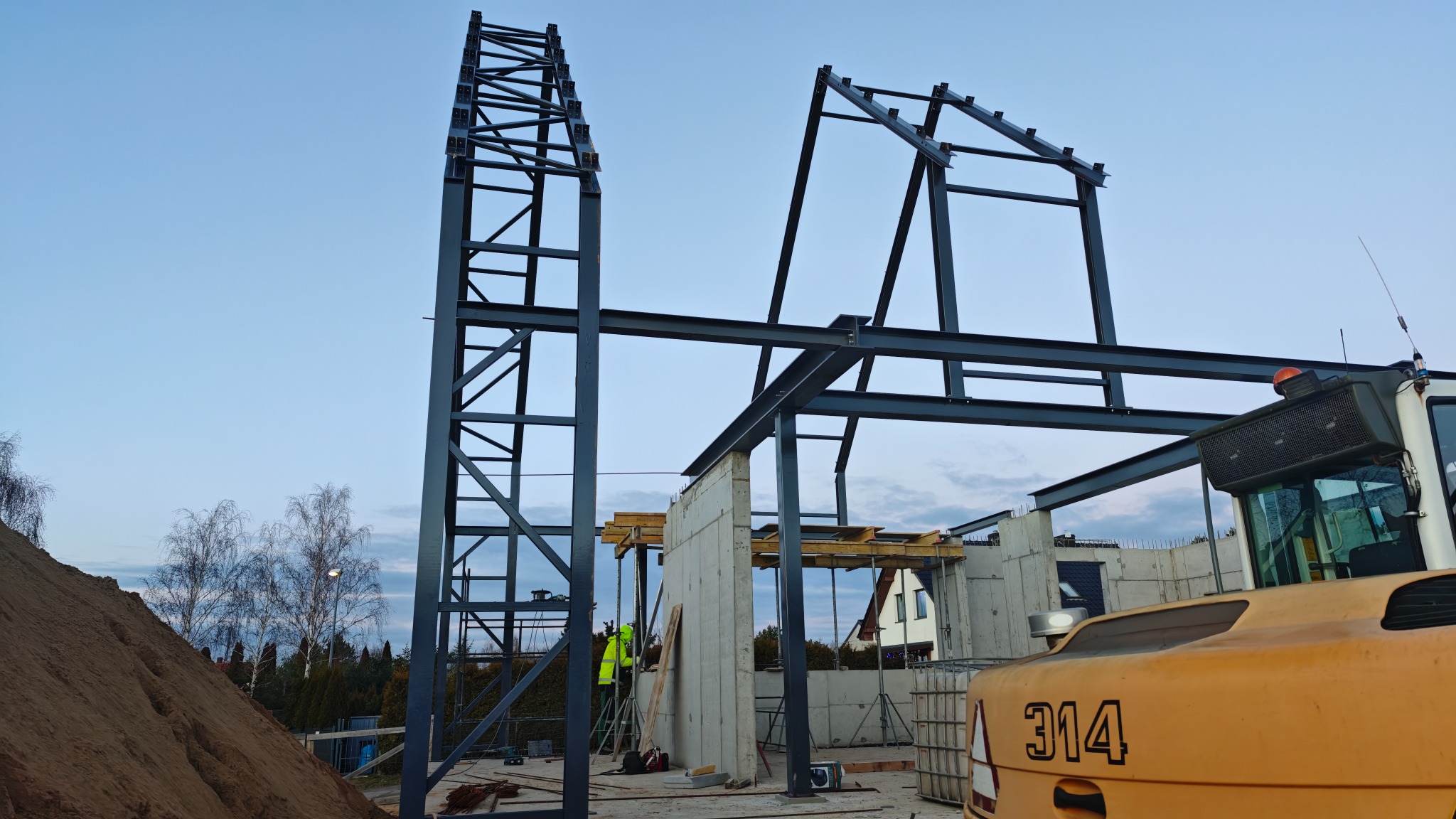 Konstrukcja budynku żelbetowego w trakcie montażu, widoczny szkielet stalowy i fragment ściany betonowej. Na pierwszym planie fragment koparki.