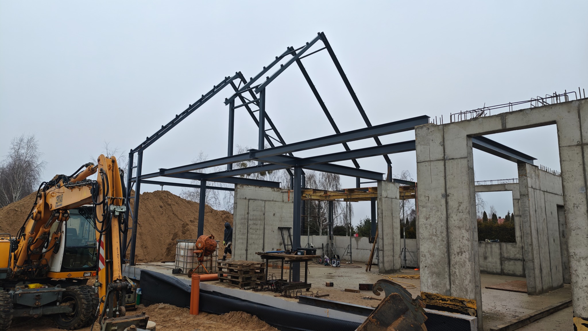 Budowa budynku żelbetowego: szkielet stalowy dachu i betonowe ściany zbrojone, widoczny żółty koparka i piasek. Etap wznoszenia konstrukcji, pochmurne niebo.