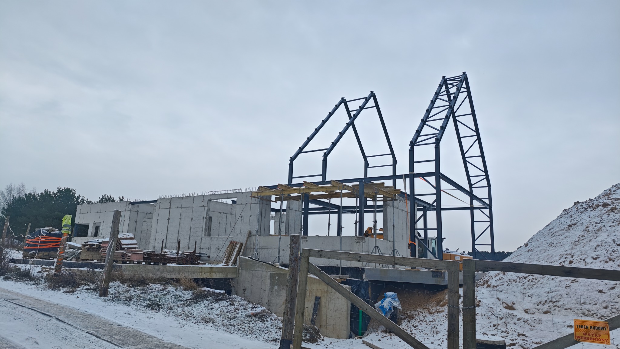 Budowa domu w zimowej scenerii: betonowe ściany, stalowa konstrukcja dachu, zasypany śniegiem teren budowy i pracownicy w odblaskowych kamizelkach.