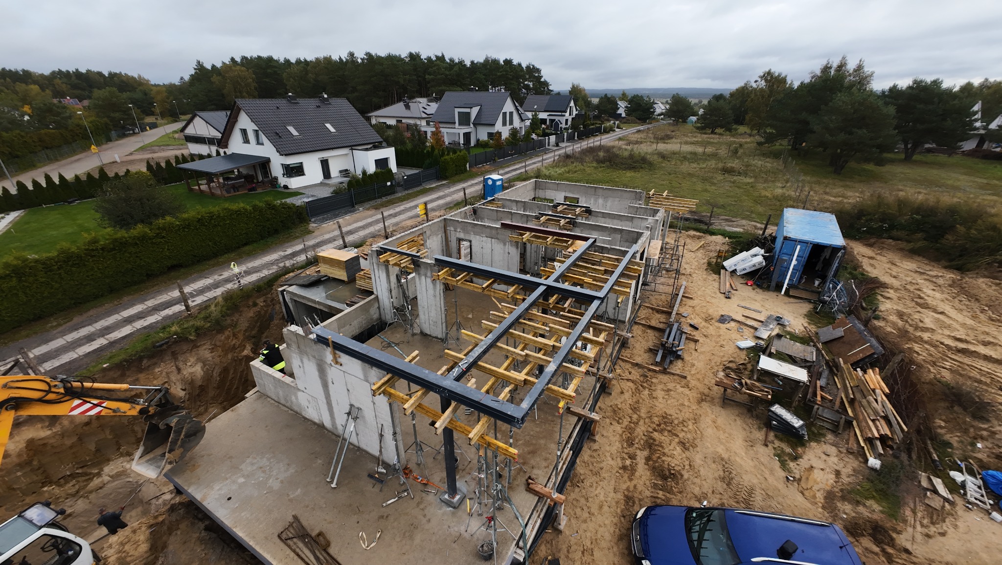 Budowa domu jednorodzinnego w stanie surowym otwartym, widok z góry na fundamenty, ściany i szalunki stropu. Na pierwszym planie wykop i koparka.