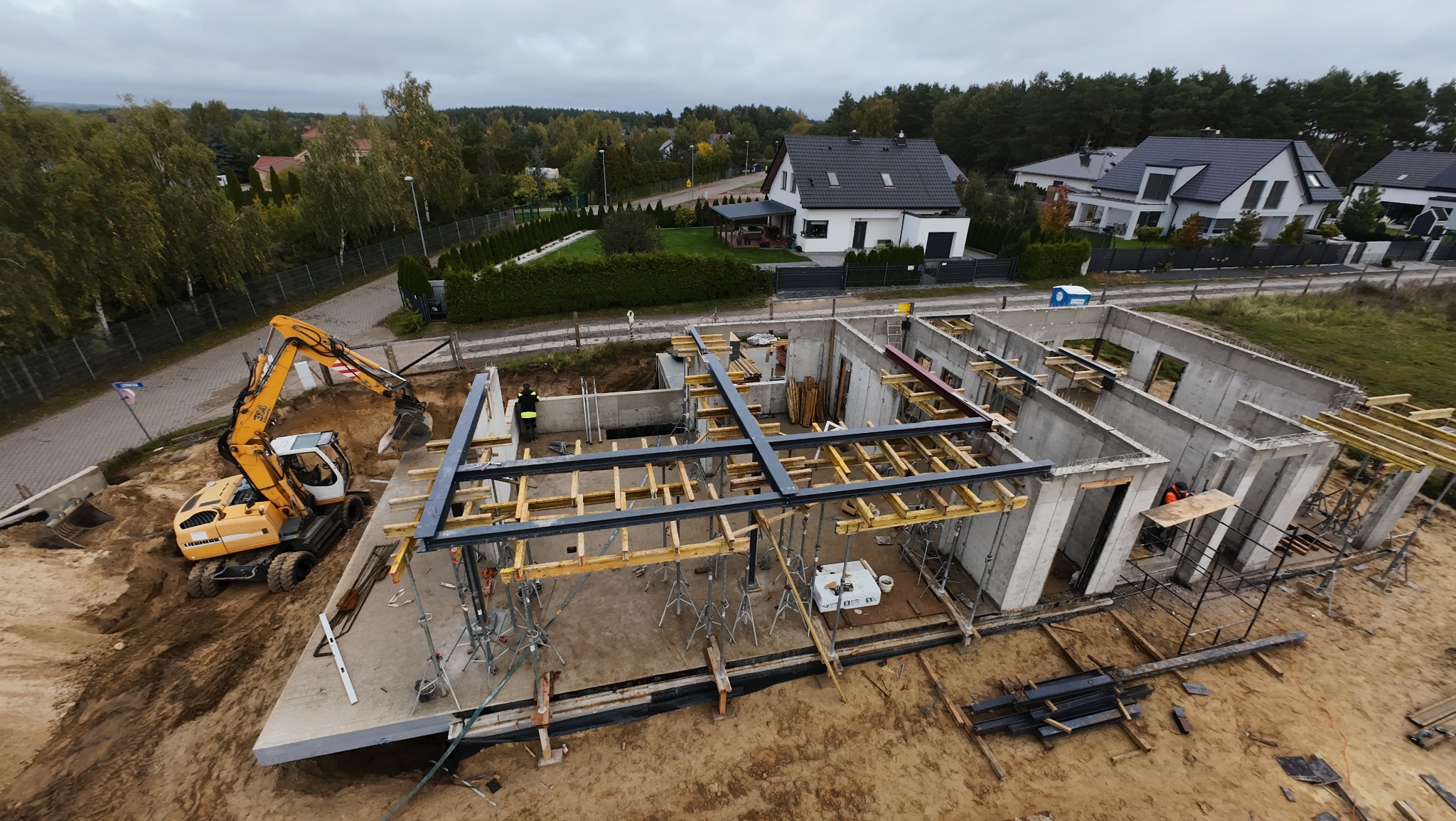 Budowa domu jednorodzinnego w trakcie realizacji: fundamenty, ściany z betonu, zbrojenie, szalunki, koparka na działce. Widok z góry na plac budowy i okoliczne domy.