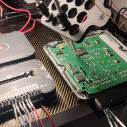 Otwarty sterownik silnika podłączony do urządzenia diagnostycznego Flex z licznymi przewodami, narzędzie precyzyjnie umieszczone nad chipem, widoczne elementy elektroniczne i złącza.