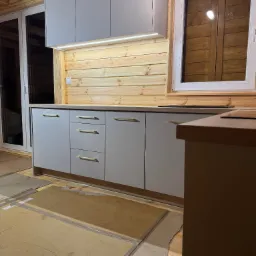 Szare szafki kuchenne z drewnianym blatem i złotymi uchwytami w domu z drewnianymi ścianami, podłoga zabezpieczona kartonem.