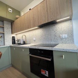 Mebelq - Nowoczesna kuchnia z drewnianymi górnymi szafkami, zielonymi dolnymi szafkami, blatem z szarego lastryko, złotą baterią i czarnym piekarnikiem.