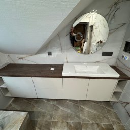 Mebelq - Biała szafka łazienkowa z ciemnobrązowym blatem pod umywalką, zamontowana w łazience na poddaszu z marmurowymi płytkami na ścianach i podłodze; okrągłe lustro odbija osobę robiącą zdjęcie.