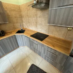 Nowoczesna kuchnia z szarymi szafkami i drewnianym blatem, czarna płyta indukcyjna, zlewozmywak i czajnik elektryczny na blacie, beżowe płytki na ścianie i jasne płytki na podłodze.