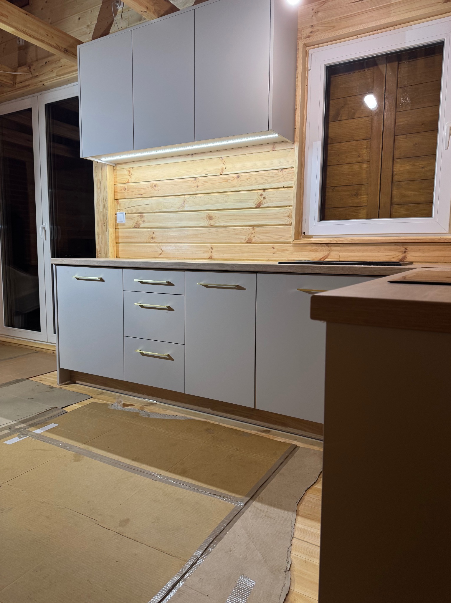 Szare szafki kuchenne z drewnianym blatem i złotymi uchwytami w domu z drewnianymi ścianami, podłoga zabezpieczona kartonem.