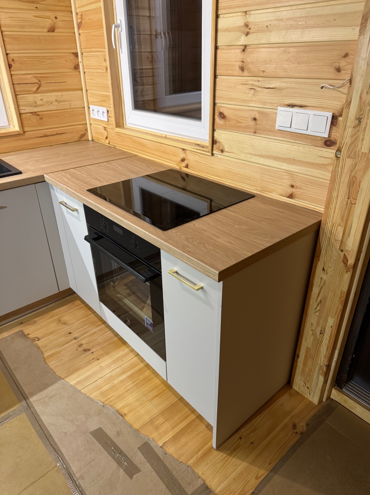 Kuchenny blat z jasnego drewna z wbudowaną płytą indukcyjną i piekarnikiem w białej zabudowie, drewniana boazeria na ścianach, widoczny fragment podłogi zabezpieczonej kartonem.