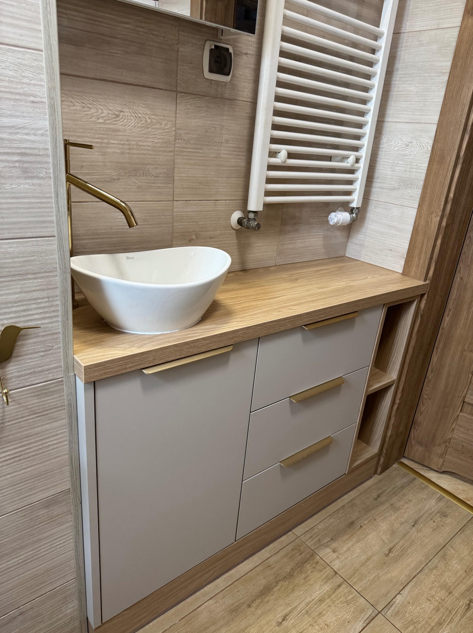 Nowoczesna łazienka z umywalką nablatową Rea, złotą armaturą i szafką z drewnianym blatem oraz grzejnikiem drabinkowym.