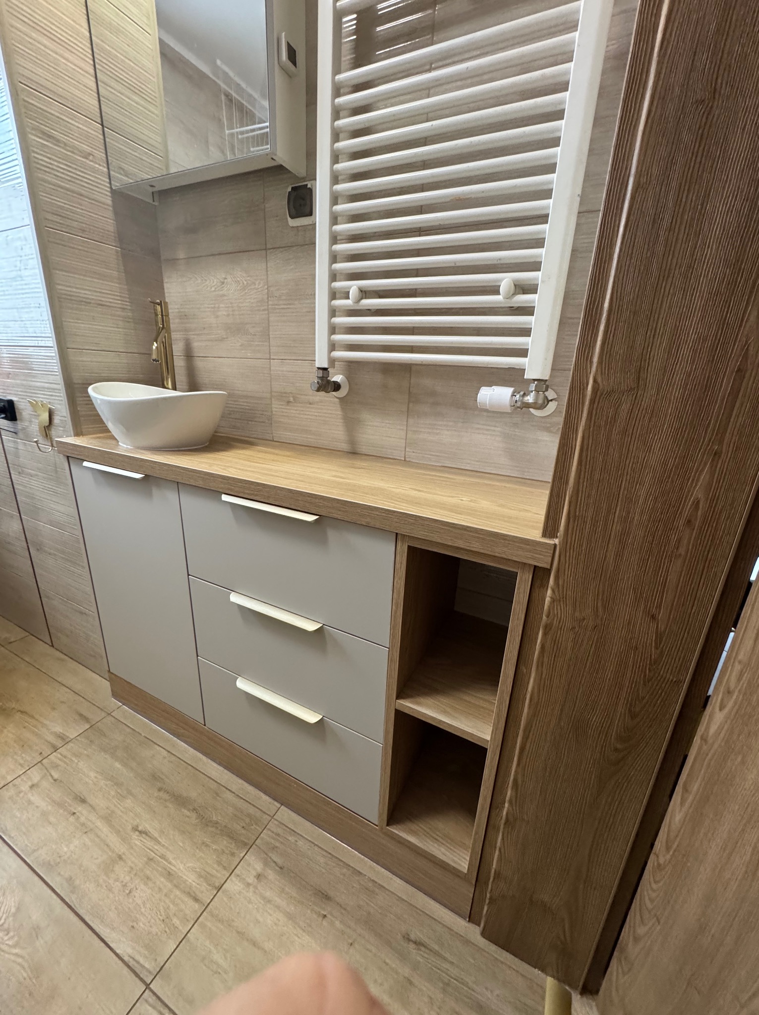 Łazienka z szarymi szafkami pod umywalką, drewnianym blatem i grzejnikiem drabinkowym na ścianie z płytek imitujących drewno.