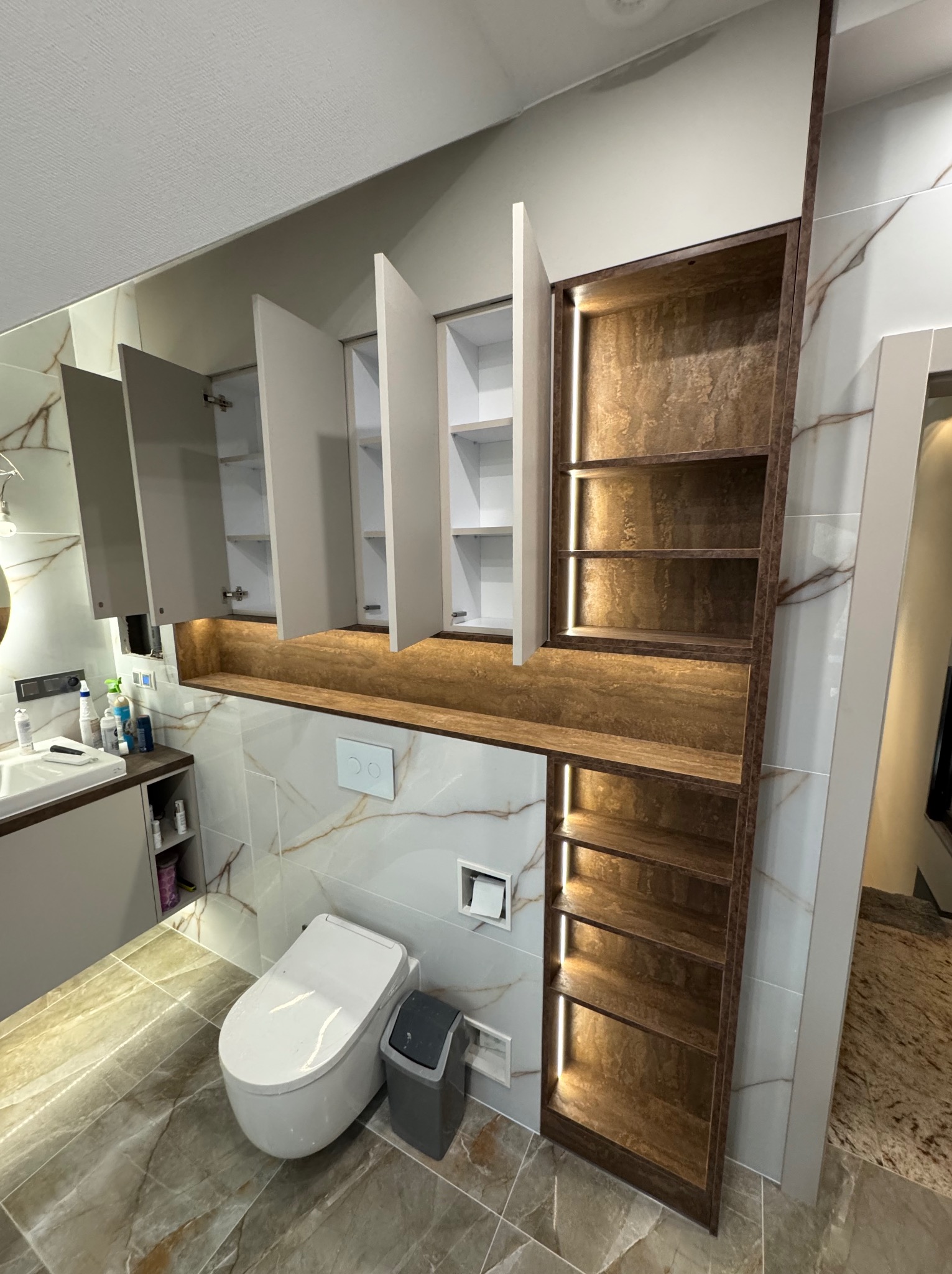 Nowoczesna łazienka z zabudową meblową: białe szafki wiszące z otwartymi drzwiczkami, podświetlane półki z teksturą drewna, umywalka z blatem i toaleta na tle marmurowych płytek.