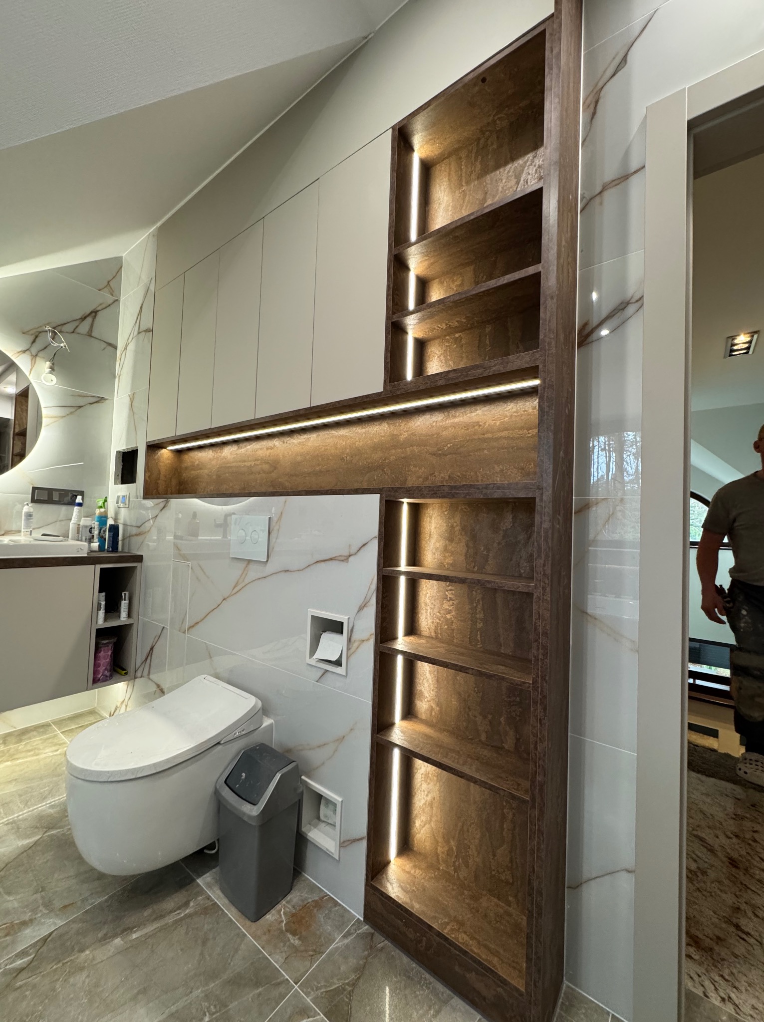 Nowoczesna łazienka z zabudową meblową: podświetlane półki z ciemnego drewna wkomponowane w jasną ścianę z marmurowym wzorem, wisząca toaleta i osoba w tle.